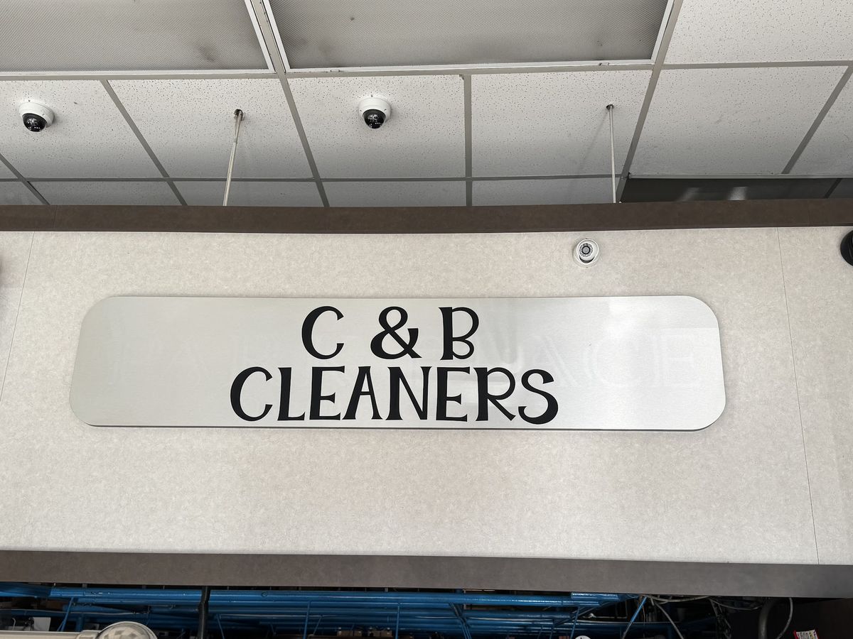 Brushed-metal C & B CLEANERS sign above the counter, with bagged garments on the conveyor below